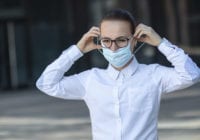 Beautiful business woman, young girl putting on medical protective mask on her face, in white shirt in glasses outdoors, healthcare at job, work, office. Coronavirus, virus, epidemic, covid-19 concept